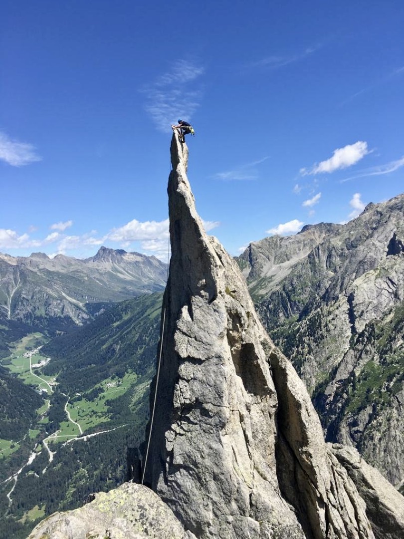 Ein sehr steiler Felsgipfel der bei blauen Himmel von dem Outdoorguide mit Kletterausrüstung erklommen wird im Hintergrund sieht man ins Tal